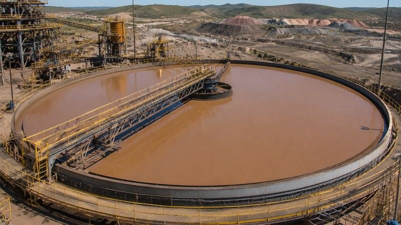 Espessador circular de grande porte em planta de processamento mineral, com água de polpa de minério e braço de raspagem central.